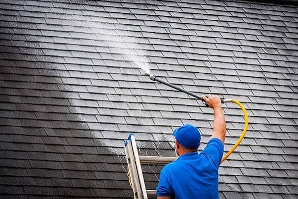 Worker in blue shirt and cap pressure washing a dark shingle roof from a ladder
