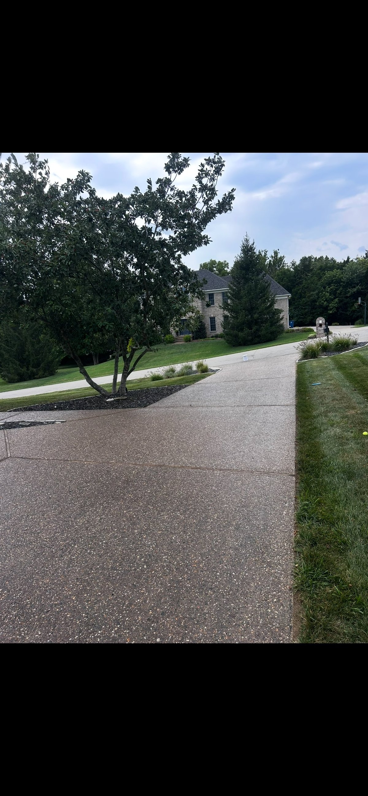 Residential driveway with gravel leading to a two-story stone house surrounded by mature trees and manicured landscaping on a sunny day