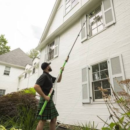 Man using a long-handled pole tool to clean gutters on a white brick residential house
