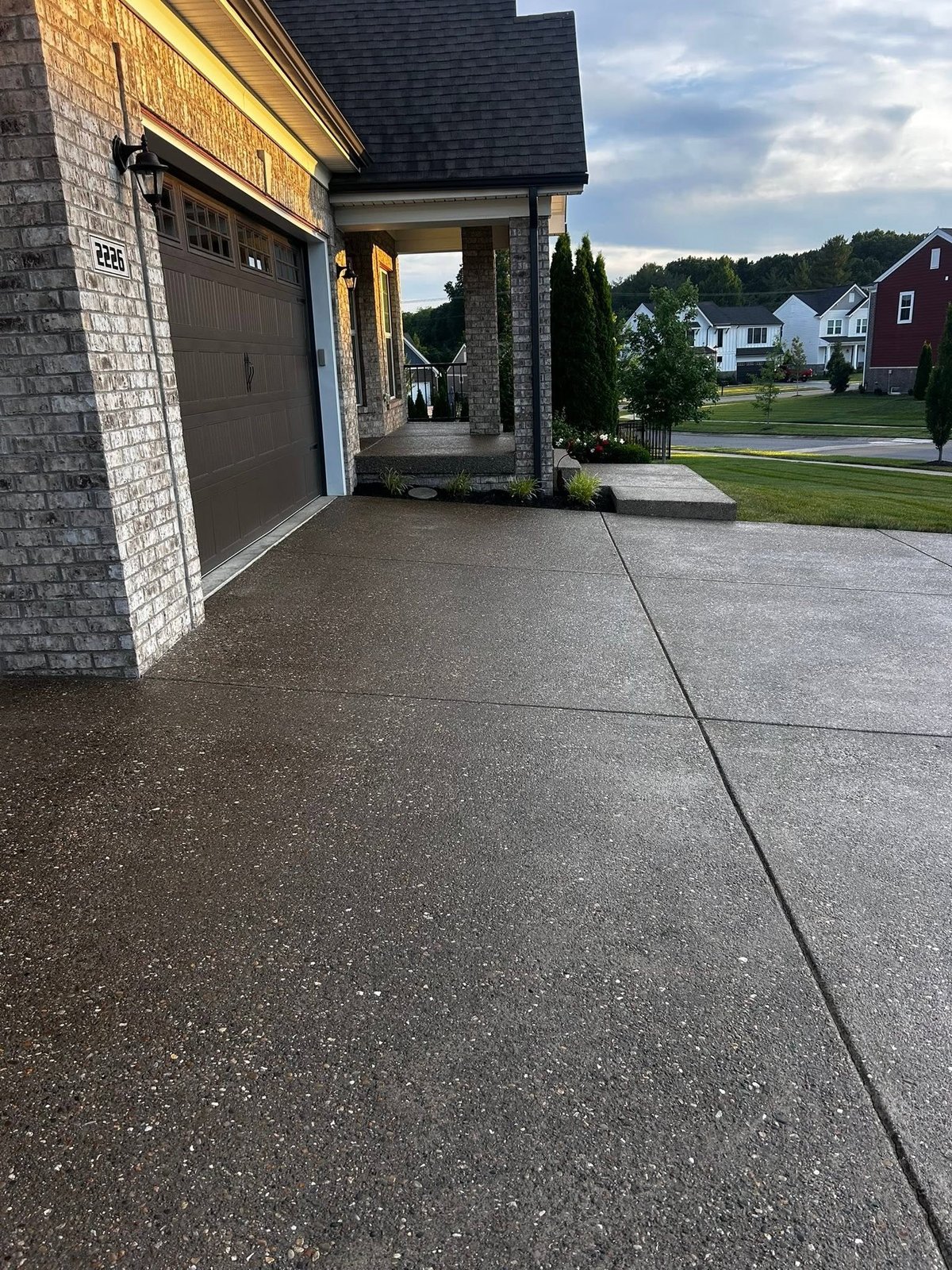 Modern brick home with attached garage and covered entryway, viewed from the driveway with freshly sealed exposed aggregate surface and neighborhood houses visible in background